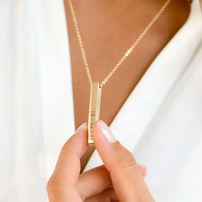 Gold necklace with a vertical bar pendant held by a hand against a white background