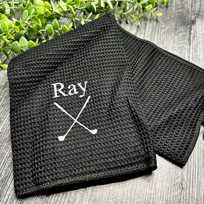 Black textured towel with personalise embroidered name and crossed golf clubs on a wooden surface with greenery.
